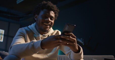 Relaxed black freelancer chatting with friends on his smartphone via texts, sitting on the couch....