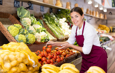 Smiling young saleswoman working in eco products store, standing near shelves with fresh vegetables and fruits, offering ripe red tomatoes