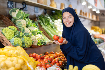 Portrait of positive young female shopper dressed in traditional muslim abaya and khimar making purchases in supermarket, choosing fresh ripe tomatoes on vegetable shelves