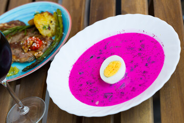Refreshing cold beetroot soup served with baked vegetables and glass of red wine on wooden background..