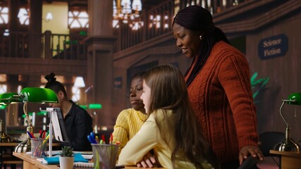 Elementary school pupils and tutor attend a live online course lesson from the library desk, connected via webcam to other classmates in a mentoring session. Virtual webinar discussion. Camera B.