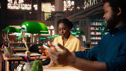 African american scholars reading course notes and comparing information at the library, studying for a test together using academic database for research. Educational materials. Camera A.