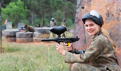 Fototapeta premium Young smiling woman paintball player in camouflage posing with gun outdoors