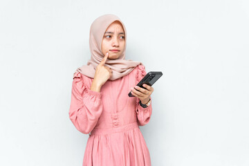Pensive beautiful Asian woman in pink dress and hijab holding mobile phone looks seriously thinking...