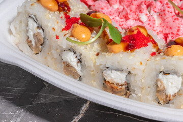 A detailed view of sushi rolls with pink creamy topping, tobiko, spicy sauce, and fresh garnish. The sushi is presented in a takeout box.