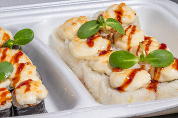 A detailed shot of baked sushi rolls covered with a melted creamy topping, teriyaki drizzle, and fresh garnish in a takeout container.
