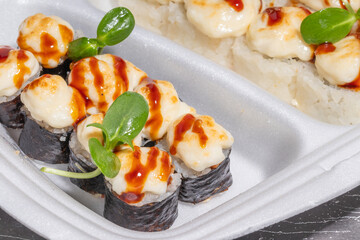 A selection of sushi rolls in a takeout box, featuring baked sushi topped with cheese-like sauce and a separate section of tobiko-covered rolls.