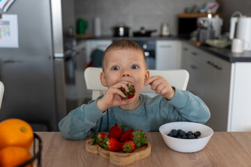 Excited little boy sitting at kitchen table, eagerly eating fresh strawberry with wide eyes and playful expression. Concept of childhood joy, healthy eating, and natural nutrition in home setting.