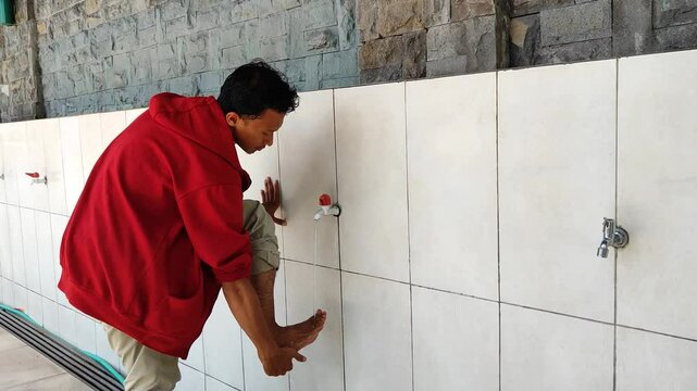 An Asian Muslim man wearing a red jacket is performing ablution (cleansing oneself with water) before praying.