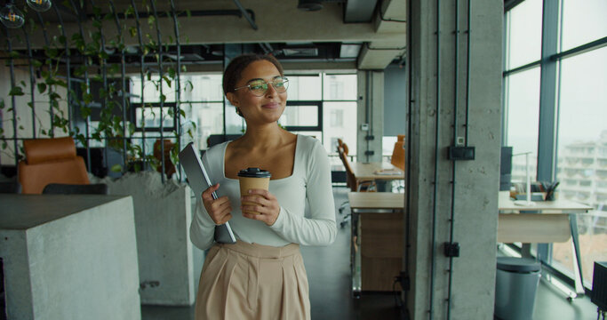 young professional woman with glasses and coffee cup walking confidently in a modern office, holding a laptop and smiling while looking out large glass windows with natural lighting