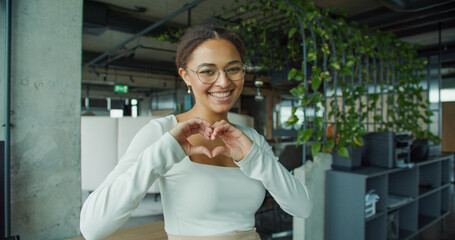 Cheerful young woman with glasses and curly hair smiling joyfully while making a heart shape with her hands, standing in a modern indoor office space with green plants and bright lighting