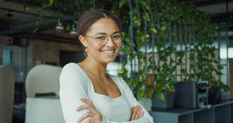 A confident, smiling young professional woman with glasses and arms crossed, standing in a modern,...