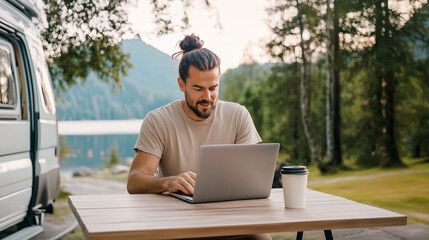 A man freelancer sits at table and works on his laptop near a travel van in the backdrop of beautiful nature during his weekend in the wilderness. Remote work during a trip.
