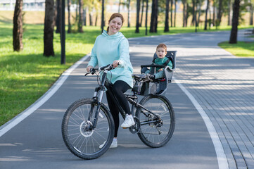 Fototapeta premium Caucasian woman riding a bicycle with her toddler son sitting behind her in a child seat. 