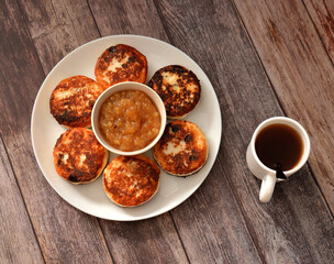 A plate with some fried cheesecakes and apple jam on a wooden table, a cup of tea with a spoon nearby.