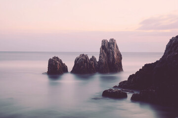 Sunset over the ocean with waves crashing in front of a rocky outcropping