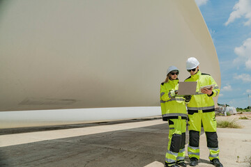 Two engineers in high visibility gear use a laptop to work in front of a large wind turbine blade...