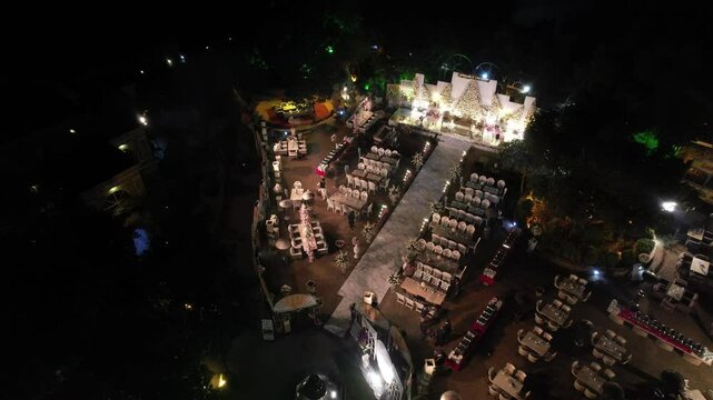Nighttime top-down zoom-out aerial drone shot of outdoor wedding reception with elegant floral stage decor, lavish tables, radiant marble-like aisle under vibrant lights&mdash;upscale events, celebrations.