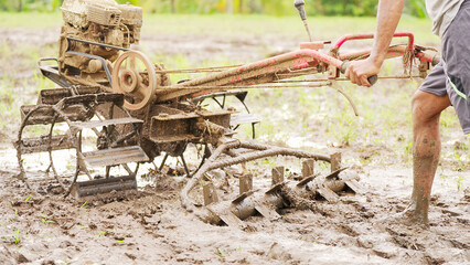 A farmer using a harrow attached to a power tiller in a muddy paddy field for rice cultivation in...