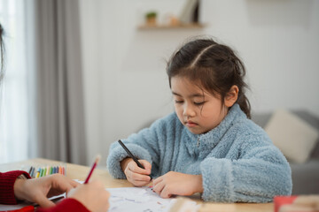 Asian Young girl in cozy blue sweater drawing with colored pencils at a wooden table in a bright home environment with focus and creativity