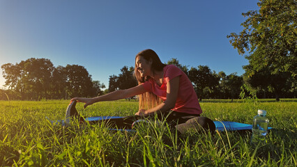 Young woman exercising on a sports yoga mat on the grass in a park during summertime sunset time.
