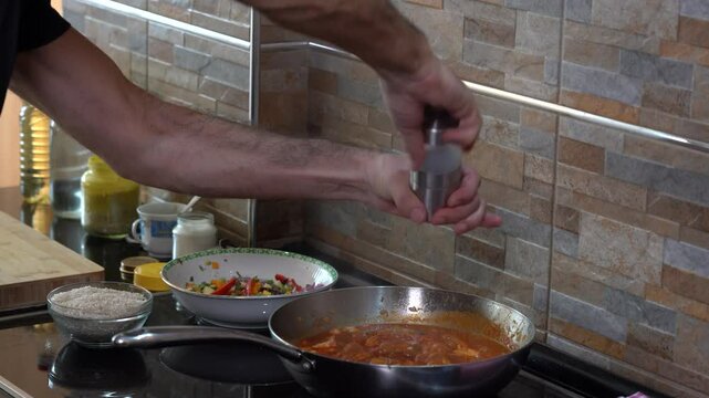 Man Salts and Adds Spices to a Meat and Vegetable Dish Cooked in Tomato Sauce. Closeup view of a frying pan on an induction plate in which a healthy meal is being cooked. Healthy Eating. Home Cooking.