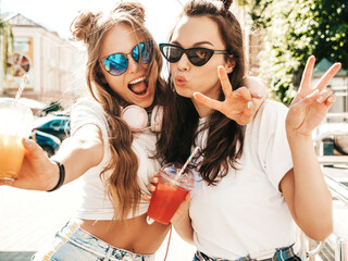 Obraz premium Two young beautiful smiling hipster female in trendy summer clothes. Carefree women posing outdoors. Positive models holding and drinking fresh cocktail smoothie drink in plastic cup with straw