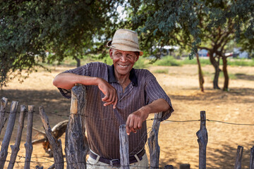 african village, old smiling man by the wooden fence in the yard, grandfather wearing a hat, sunny...