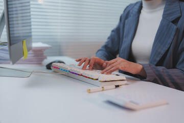 Businesswoman sits at her desk in a modern office, diligently typing on a keyboard while working on her computer, surrounded by paperwork and office supplies, focused on her tasks