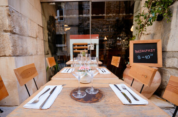 Outdoor dining setup with wooden tables and chairs, featuring neatly arranged cutlery and glasses. A chalkboard sign advertises a Dalmatian breakfast special. Stone walls and plants add charm.