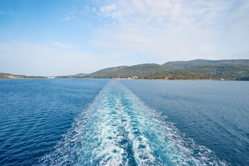View from a boat showing a wake trail on a calm sea, with a distant coastal city and mountains under a cloudy sky.