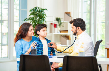 Male doctor checking boy’s symptoms with stethoscope as mother watches in hospital clinic setting