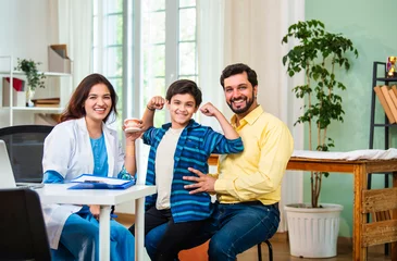 Foto auf Acrylglas Zahnärzte Happy Indian boy flexing muscles to show strong teeth while dentist and father smile in clinic  © StockImageFactory