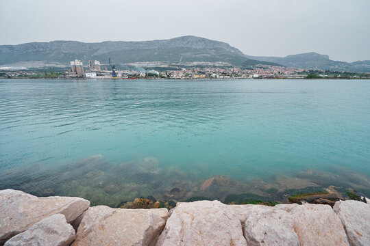 A scenic view of a calm turquoise sea with a rocky shoreline in the foreground and a distant town and mountains in the background under a cloudy sky.