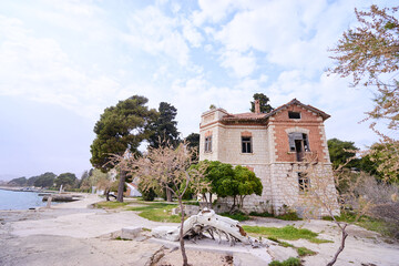 An abandoned, dilapidated stone house by the seaside, surrounded by trees and a cloudy sky.