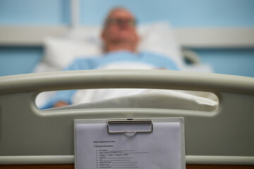 Senior patient resting on hospital bed with medical records clipboard visible in foreground, giving...