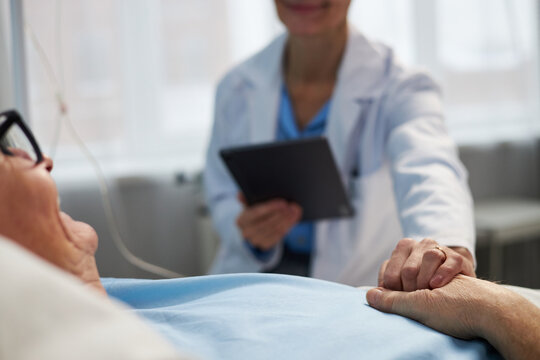 Doctor standing by patient bedside holding patient's hand, providing comfort and support during medical examination. Patient lying in bed receiving care without visible stress