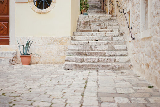 A narrow stone staircase leads up through a quaint alleyway, flanked by rustic stone walls. The steps are uneven and weathered, suggesting age.  - Powered by Adobe