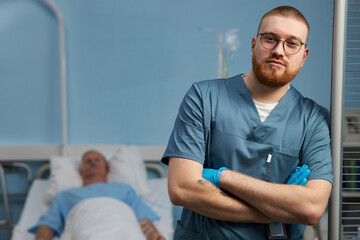 Obraz premium Doctor standing with arms crossed in hospital room by patient bedside wearing medical scrubs. Patient resting on hospital bed covered with a blanket in the background