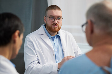 Portrait of young male doctor wearing a white coat, talking with colleagues during a meeting....