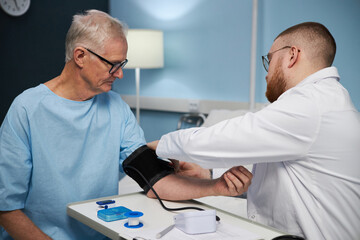 Fototapeta premium Medical professional checking blood pressure of senior Caucasian male patient in clinical setting with various medical devices present