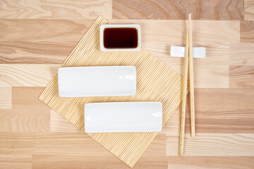 A refined sushi table setting on a light wooden surface, featuring two rectangular white ceramic plates, a small dish of soy sauce, a pair of bamboo chopsticks