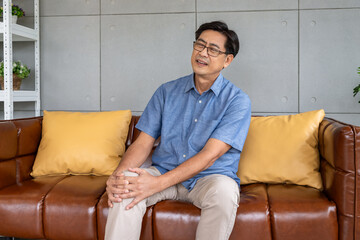 Asian elderly man suffering from knee pain while sitting on a brown leather sofa in a modern living...