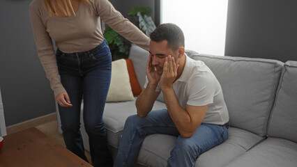 Woman comforting man sitting on sofa in living room, highlighting emotional support in a cozy apartment setting with plants and cushions.