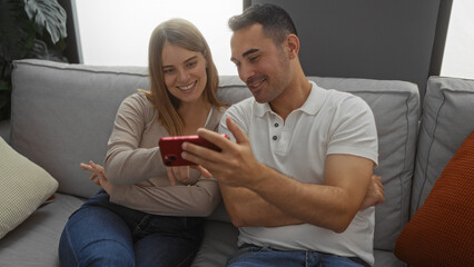 Couple sitting together on a couch indoors, man and woman smiling while looking at a smartphone, showcasing love and connection in a cozy living room setting.