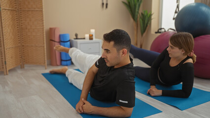 Obraz premium Man and woman practicing yoga on mats in gym with fitness equipment, emphasizing relaxation and exercise in a calm indoor environment.