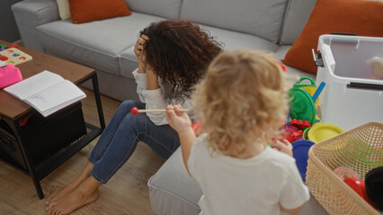 Woman sitting tired on living room floor while child plays indoors with colorful toys showing...