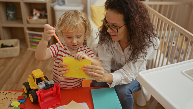Mother and son bonding indoors, creating art in cozy bedroom with toys around, highlighting loving family moments in a home setting with woman assisting child playfully.