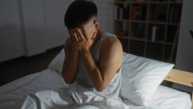Young man sitting on a bed with his hands covering his face in a dimly lit bedroom, appearing stressed or upset, surrounded by a cozy home atmosphere.