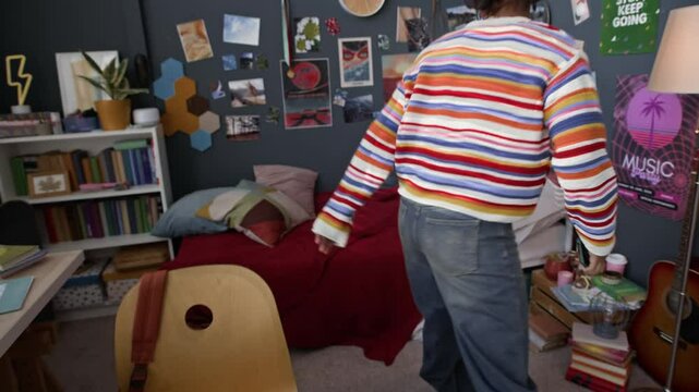 Handheld tracking shot of African American gen z girl wearing headphones completing homework and moving on bed texting messages and checking social media on mobile phone in room with many posters
